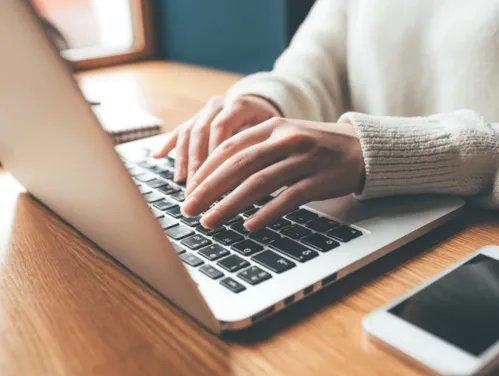 Woman types on laptop with her cell phone nearby.