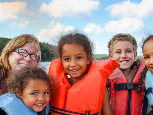 a woman and four small kids wearing life jackets smile at the camera in a boat.