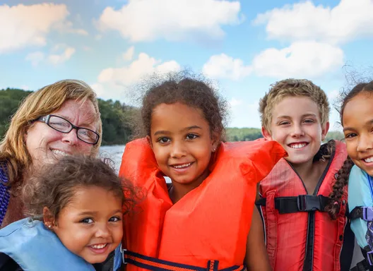 a woman and four small kids wearing life jackets smile at the camera in a boat.